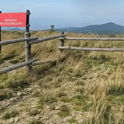 Czerwony znak informuje o wejściu na teren Śnieżnickiego Parku Krajobrazowego. Drewniane ogrodzenie i trawiasty, kamienisty teren prowadzą w głąb gór.
