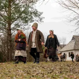 Ludzie w strojach z epoki, przypominający mieszkańców wsi, idą przez trawiasty teren. W tle widać drewniane domy i drzewa. Wśród nich są krużewniki.